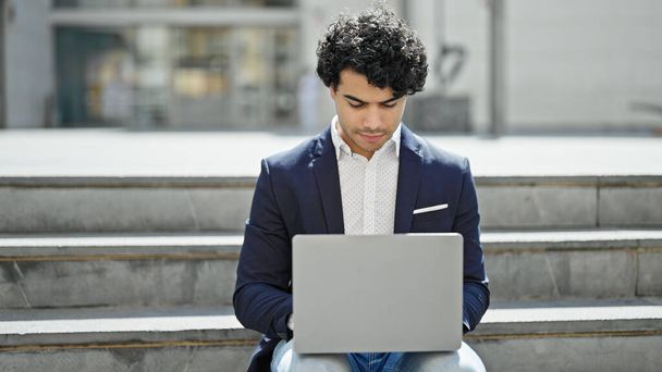 Молодий латинський бізнесмен використовує ноутбук, сидячи на підлозі на вулиці - Фото, зображення