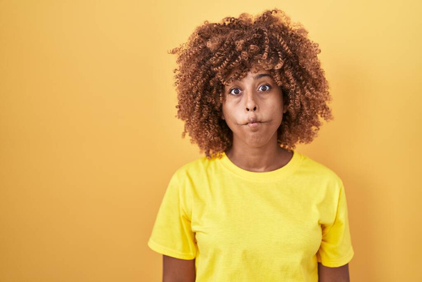 Young hispanic woman with curly hair standing over yellow background making fish face with lips, crazy and comical gesture. funny expression.  - Photo, image