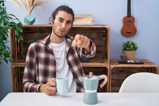 Молодий іспаномовний чоловік п'є каву від французької кавоварки, вказуючи пальцем на камеру і до вас, впевнений жест виглядає серйозно  - Фото, зображення
