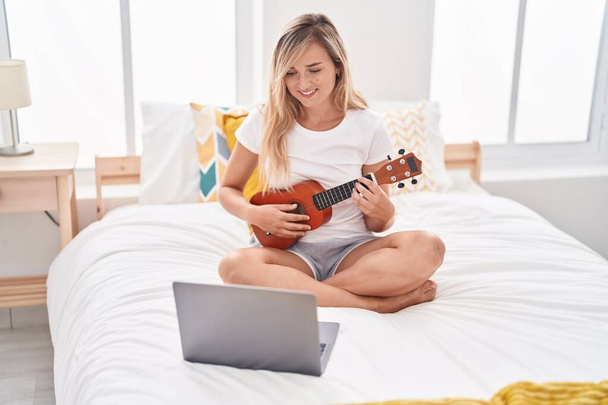 fiatal szőke nő miután online ukulele lecke ül az ágyon a hálószobában - Fotó, kép