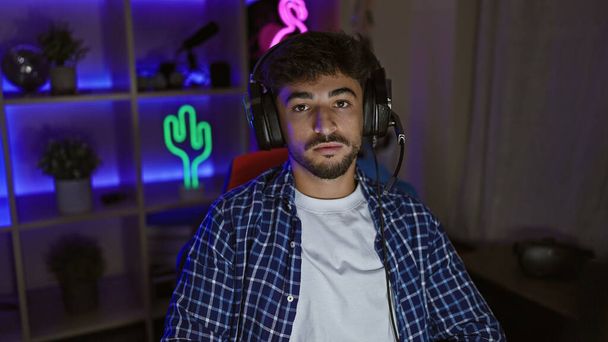 Focado no jogo, jovem árabe, um streamer sério, usando fones de ouvido em uma sala de jogos escura, absorvido em um mundo futurista de jogos virtuais - Foto, Imagem