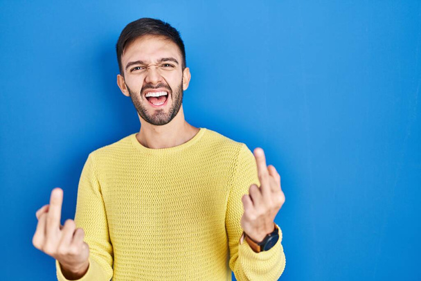 Hispanic man standing over blue background showing middle finger doing fuck you bad expression, provocation and rude attitude. screaming excited  - Фото, изображение