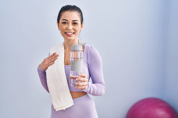 若い美しいヒスパニックの女性は,スポーツセンターで水のボトルを保持する自信を持って微笑む - 写真・画像