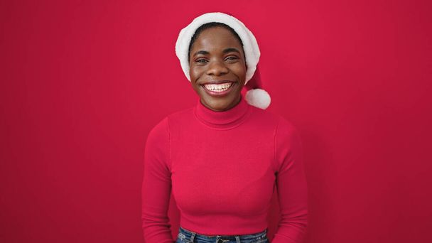 Mujer afroamericana sonriendo confiada usando sombrero de navidad sobre fondo rojo aislado - Foto, imagen