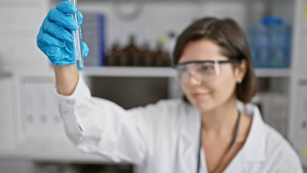 Itsevarma & kaunis nuori latino nainen tiedemies, hymyillen pitäen koeputki, kovaa työtä vilkkaassa laboratoriossa, paistattelu hehku hänen säteilevä kokeiluja. - Valokuva, kuva