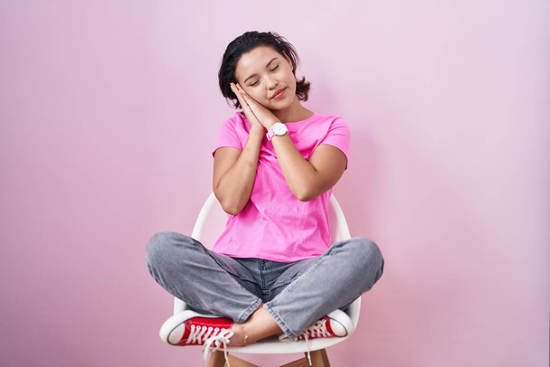 Mulher jovem hispânica sentada na cadeira sobre fundo rosa dormindo cansado sonhando e posando com as mãos juntas enquanto sorri com os olhos fechados.  - Foto, Imagem