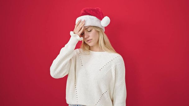 Νεαρή ξανθιά γυναίκα με χριστουγεννιάτικο καπέλο που υποφέρει από πονοκέφαλο σε απομονωμένο κόκκινο φόντο - Φωτογραφία, εικόνα