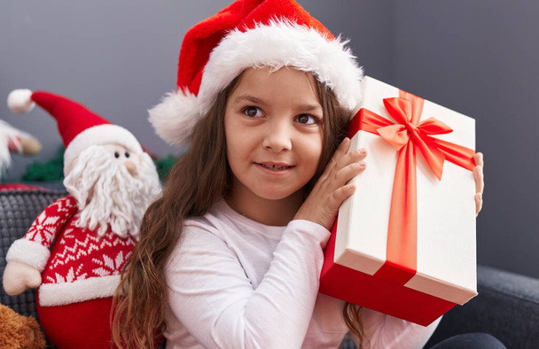 Adorabile ragazza ispanica che sente il suono del regalo di Natale seduto sul divano a casa - Foto, immagini