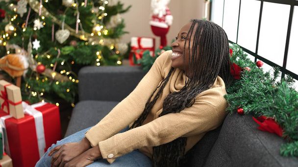 Afrikaanse vrouw glimlachen zitten door kerstboom thuis - Foto, afbeelding