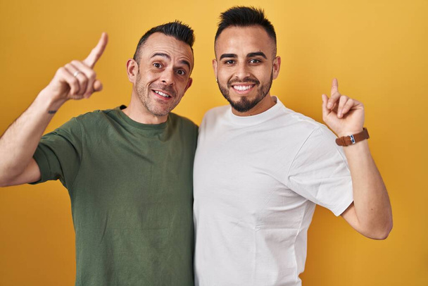 Homosexuelles Paar, das vor gelbem Hintergrund steht, erstaunt und überrascht lächelt und mit erhobenen Fingern und Armen nach oben zeigt.  - Foto, Bild