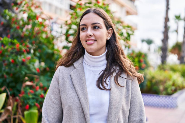 Jonge mooie Spaanse vrouw glimlachend zelfverzekerd kijkend naar de zijkant in het park - Foto, afbeelding