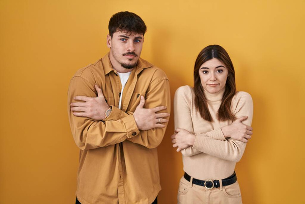 Young hispanic couple standing over yellow background shaking and freezing for winter cold with sad and shock expression on face  - Photo, Image
