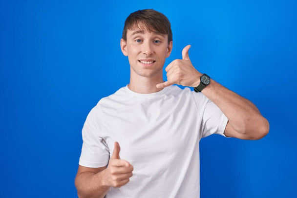 Caucasian blond man standing over blue background smiling doing talking on the telephone gesture and pointing to you. call me.  - Photo, Image