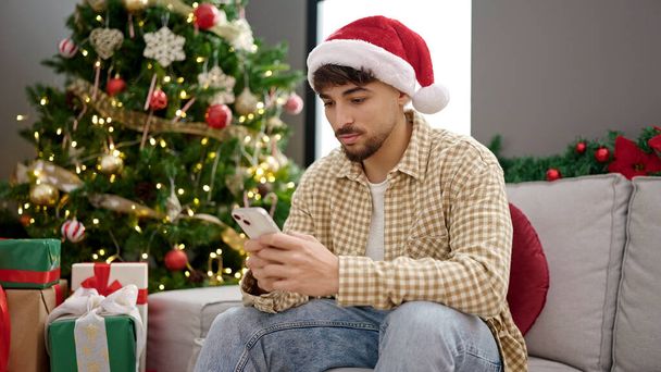 Giovane arabo utilizzando smartphone seduto vicino all'albero di Natale a casa - Foto, immagini