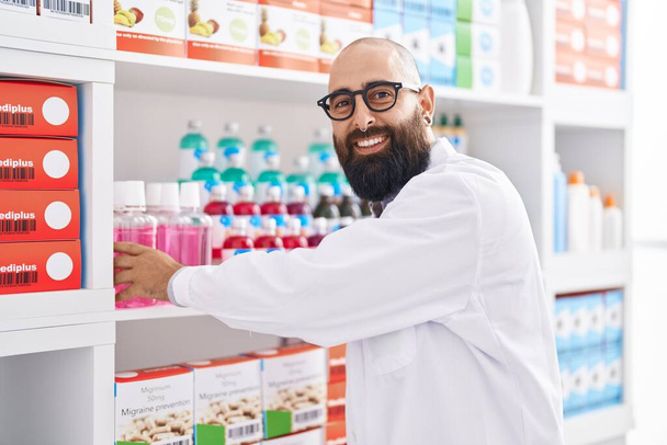 Fiatal kopasz férfi gyógyszerész mosolygós magabiztos gazdaság gyógyszert polcon gyógyszertár - Fotó, kép