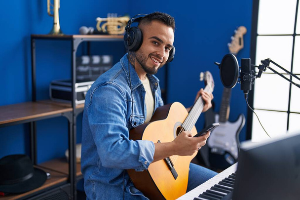Giovane artista uomo ispanico utilizzando smartphone suonare la chitarra classica in studio di musica - Foto, immagini
