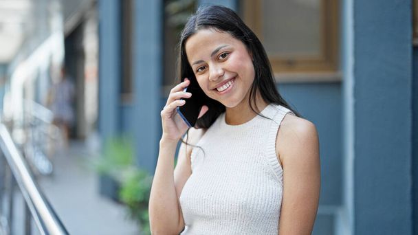 Junge schöne hispanische Frau lächelt selbstbewusst und spricht auf der Straße mit dem Smartphone - Foto, Bild