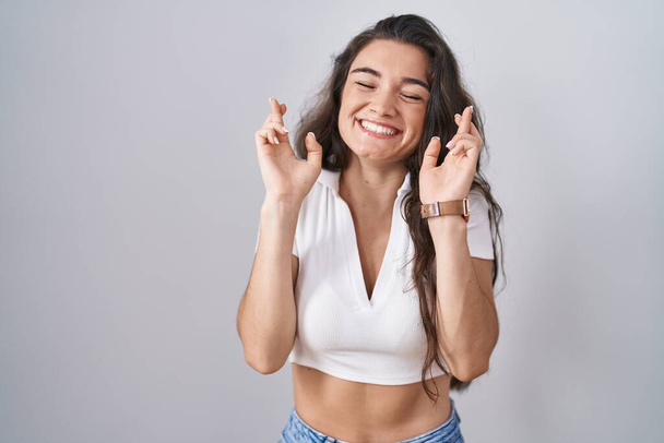 Young teenager girl standing over white background gesturing finger crossed smiling with hope and eyes closed. luck and superstitious concept.  - Photo, Image