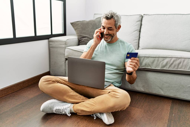 Седовласый мужчина средних лет разговаривает по смартфону, держа кредитку на полу дома - Фото, изображение