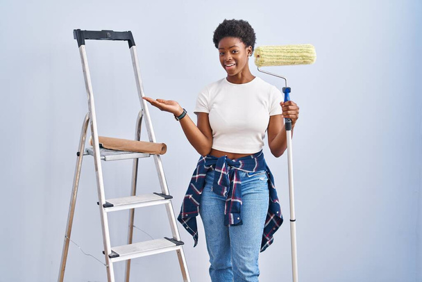 Mujer afroamericana sosteniendo rodillo pintor señalando a un lado con las manos abiertas palmeras mostrando espacio de copia, presentando anuncio sonriendo emocionado feliz  - Foto, imagen
