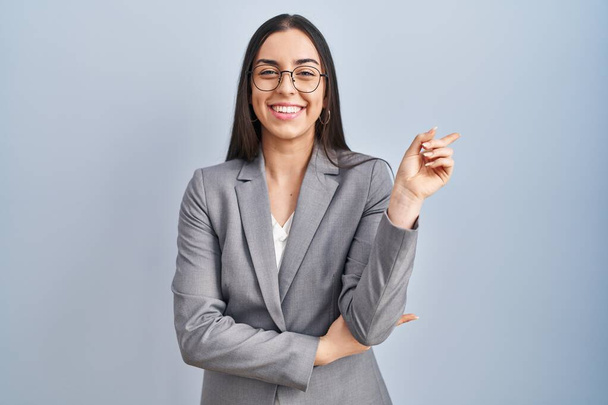 Femme d'affaires hispanique portant des lunettes avec un grand sourire sur le visage, pointant avec la main et le doigt vers le côté regardant la caméra.  - Photo, image