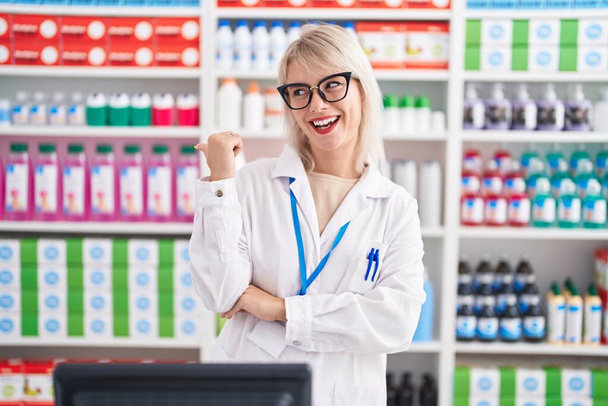 Jonge blanke vrouw werkt bij apotheek drogisterij glimlachend met een vrolijk gezicht en wijzend naar de zijkant met duim omhoog.  - Foto, afbeelding
