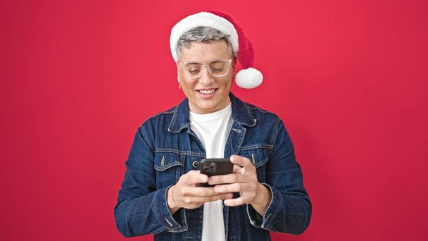 Молодой кавказский мужчина в рождественской шляпе с помощью смартфона улыбается на изолированном красном фоне - Фото, изображение