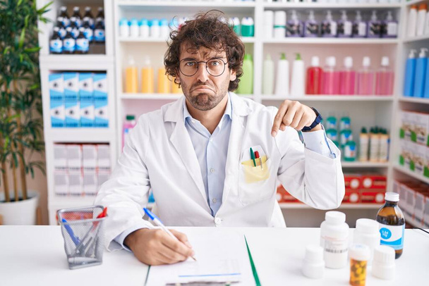 Ισπανόφωνος νεαρός άνδρας που εργάζεται στο φαρμακείο δείχνει λυπημένος και αναστατωμένος, υποδεικνύοντας κατεύθυνση με τα δάχτυλα, δυστυχισμένος και καταθλιπτικός.  - Φωτογραφία, εικόνα