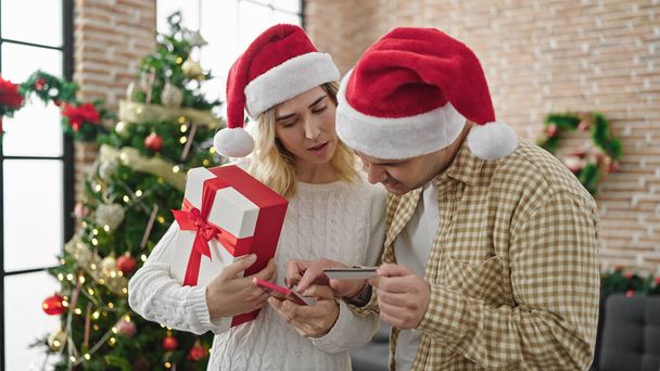 Άνδρας και γυναίκα ζευγάρι κρατώντας χριστουγεννιάτικο δώρο ψώνια με smartphone και πιστωτική κάρτα στο σπίτι - Φωτογραφία, εικόνα