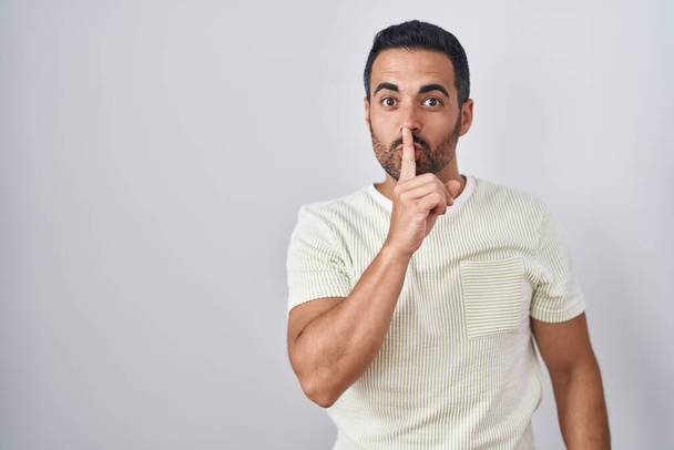 Homme hispanique avec la barbe debout sur fond isolé demandant à être calme avec le doigt sur les lèvres. silence et concept secret.  - Photo, image