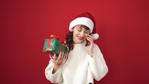 Młoda biała kobieta uśmiechnięta w świątecznym kapeluszu z prezentem rozmawiająca przez telefon przez odizolowane czerwone tło - Zdjęcie, obraz