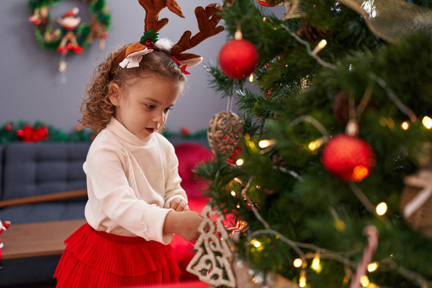Entzückendes blondes Mädchen mit Rentierohren schmückt zu Hause den Weihnachtsbaum - Foto, Bild