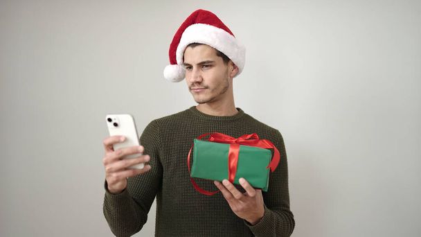 Молодой латиноамериканец в рождественской шляпе с помощью смартфона на изолированном белом фоне - Фото, изображение