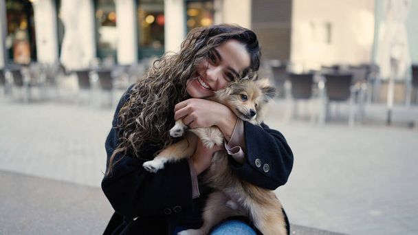 Köpeğiyle gülümseyen genç İspanyol kadın sokakta bankta oturuyor. - Fotoğraf, Görsel