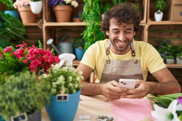 Jonge Spaanse man bloemist glimlacht vol vertrouwen met behulp van smartphone bij de bloemenwinkel - Foto, afbeelding
