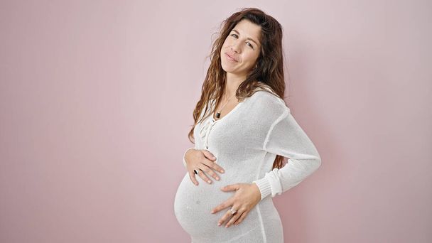 Jovem grávida sorrindo confiante tocando barriga sobre fundo rosa isolado - Foto, Imagem