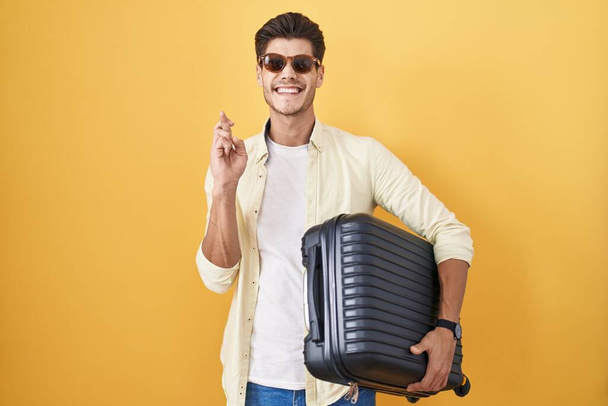 Młody Latynos trzymający walizkę na wakacjach gestykulujący palec skrzyżowany uśmiechnięty z nadzieją i zamkniętymi oczami. szczęście i przesądna koncepcja.  - Zdjęcie, obraz