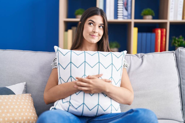 Mujer hispana abrazando almohada sentada en el sofá sonriendo mirando hacia un lado y mirando hacia otro pensando.  - Foto, Imagen