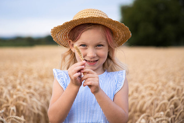 小麦畑で帽子をかぶっている笑顔の子供の金髪の少女. - 写真・画像