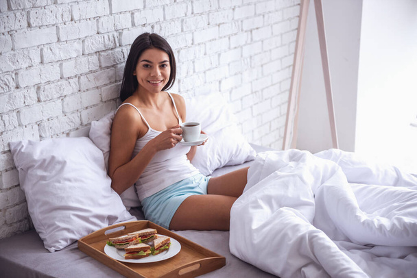 Доброе утро! Счастливая молодая красивая женщина завтракает, сидя дома на кровати
 - Фото, изображение
