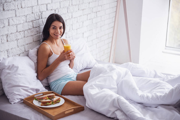 Καλημέρα! Ευτυχισμένος νεαρή όμορφη γυναίκα τρώει πρωινό, ενώ κάθεται στο κρεβάτι στο σπίτι - Φωτογραφία, εικόνα