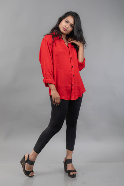 Ινδικό νεαρό θηλυκό μοντέλο σε casual φορούν σε γκρι φόντο - φωτογραφία αρχείου. Μακριά μαύρα μαλλιά μοντέλο φορώντας κόκκινο πουκάμισο και μαύρο τζιν. - Φωτογραφία, εικόνα