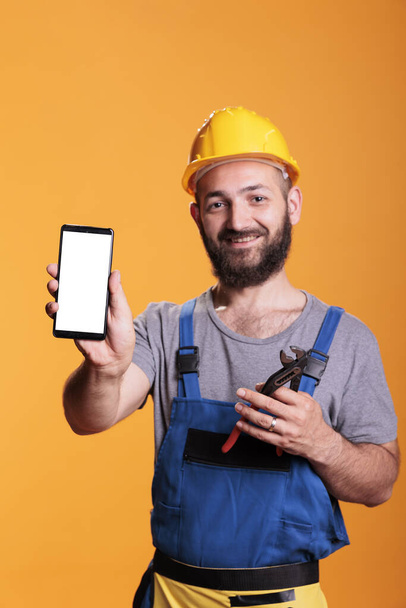 カメラの前に広告のための空のスクリーンが付いている携帯電話を握る構造労働者. スタジオショットで黄色の背景に対する硬い帽子を持つプロのビルダー. - 写真・画像