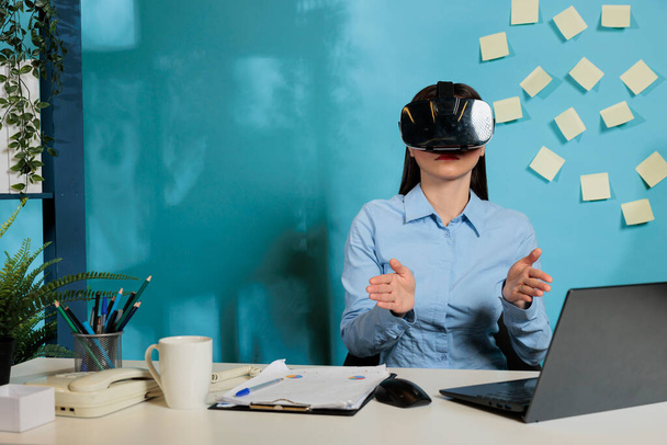Empleada corporativa con gafas de realidad virtual para visualizar proyecto de trabajo. Mujer profesional con visor multimedia inmersivo trabajando en escritorio de oficina gesticulando con las manos. - Foto, imagen