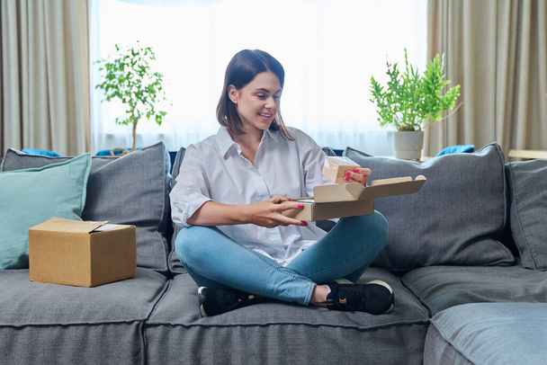 Young woman unpacking cardboard box with internet order, online purchase of cosmetic care products, sitting on sofa at home. Online shopping, delivery service concept - Foto, immagini