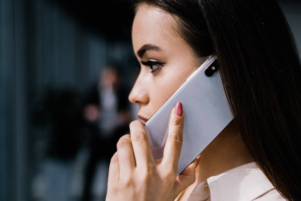 Side view of young attractive serious female in formal clothes attentively talking on modern smartphone standing at workplace looking away - Foto, afbeelding