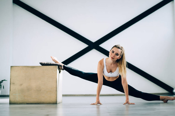 Magnificent slim determined woman in sport clothes smiling at camera while doing front split exercise leaning on wooden box and floor against black sports wall resistance bands in cozy modern gym - Фото, изображение