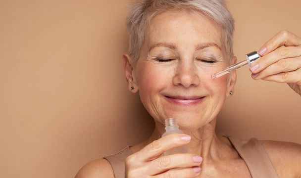 Happy senior lady applies cosmetic oil serum on face takes care of skin and smiles broadly enjoys beauty treatments. Portrait over beige background. - Photo, image