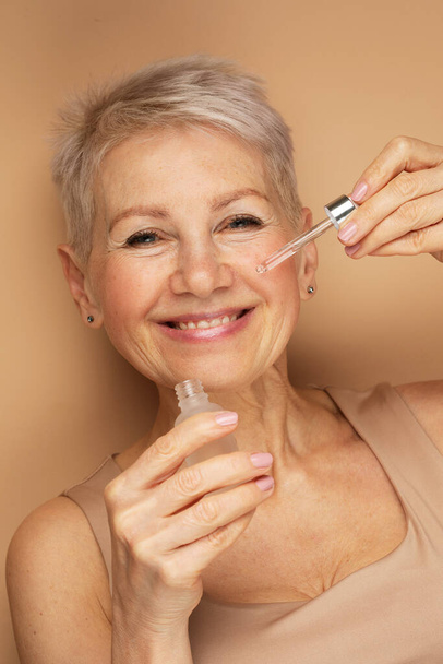 Happy senior lady applies cosmetic oil serum on face takes care of skin and smiles broadly enjoys beauty treatments. Portrait over beige background. - Foto, imagen
