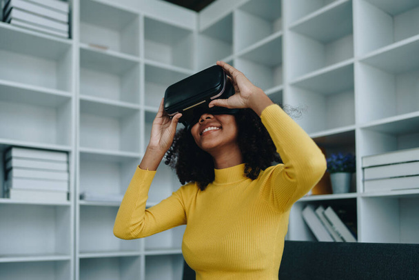 Positive African American woman teenager in futuristic VR helmet laughing and relaxing. Youthful girl is delighted with high-tech video games and virtual reality gadgets sits, Artificial Intelligence. - Photo, Image
